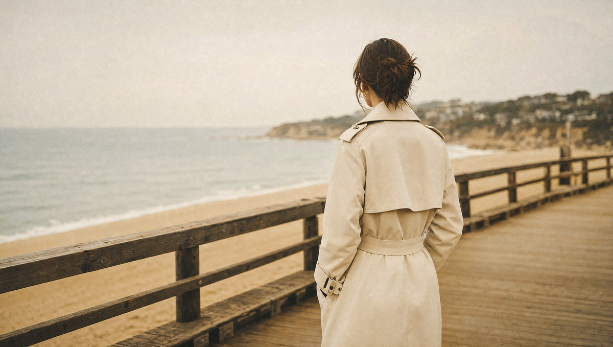 Woman in a cream trench coat on a quiet coastal boardwalk in soft morning light